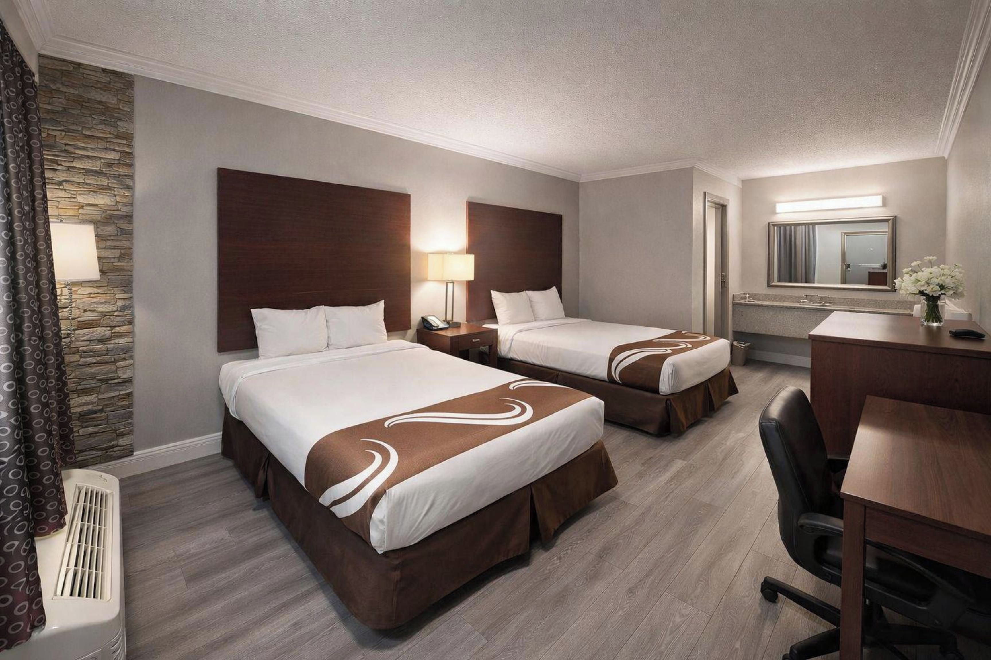 A hotel room with two beds, a desk, a chair, a mirror, and a bathroom.