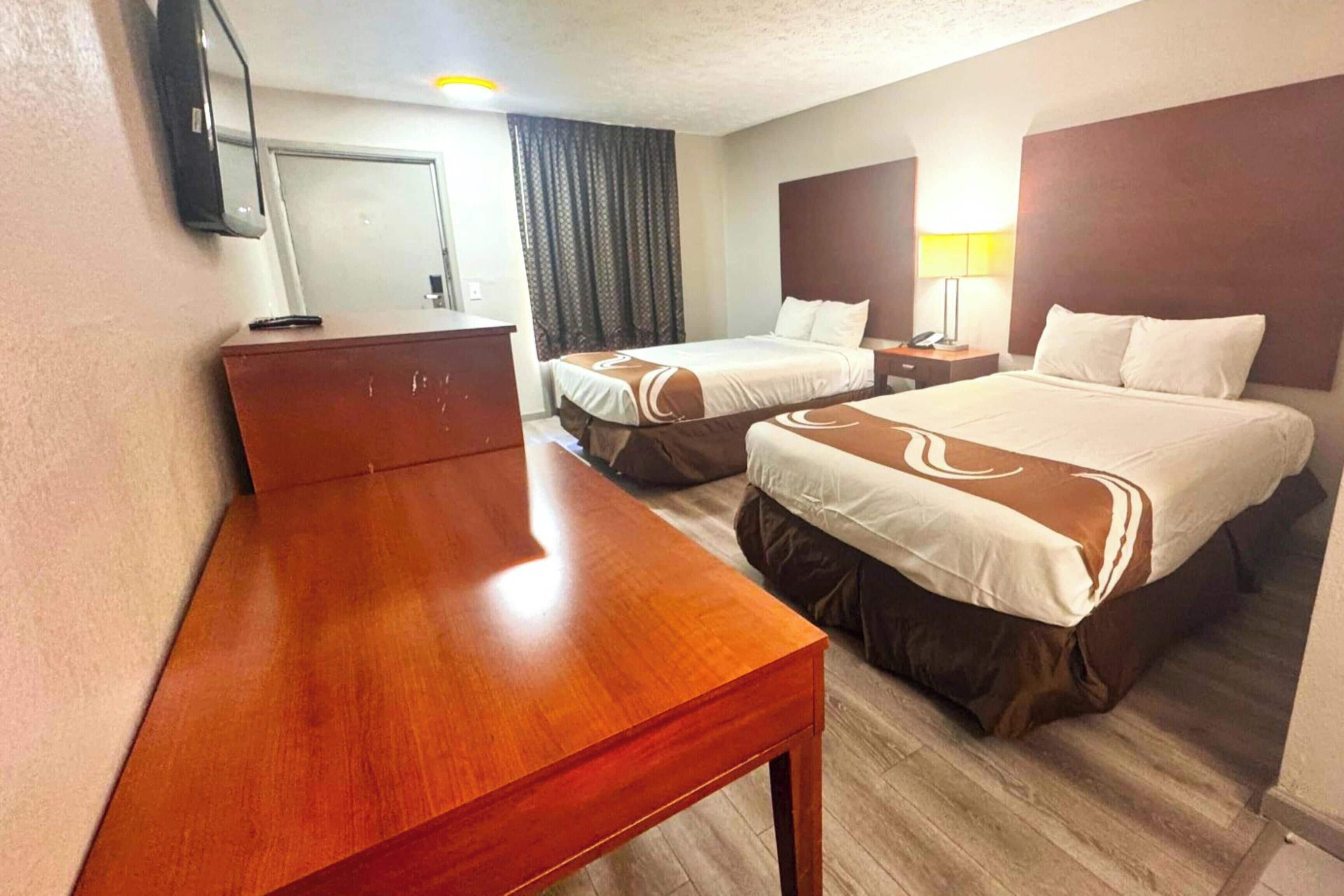A hotel room with two beds, a wooden desk, a TV, and a nightstand with a lamp.