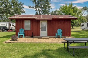 Un petit bâtiment rouge avec une porte et deux fenêtres, deux chaises turquoise, une table de pique-nique et un barbecue.