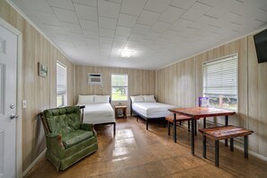 Une chambre avec deux lits, un fauteuil vert, une table en bois et une fenêtre avec des stores.