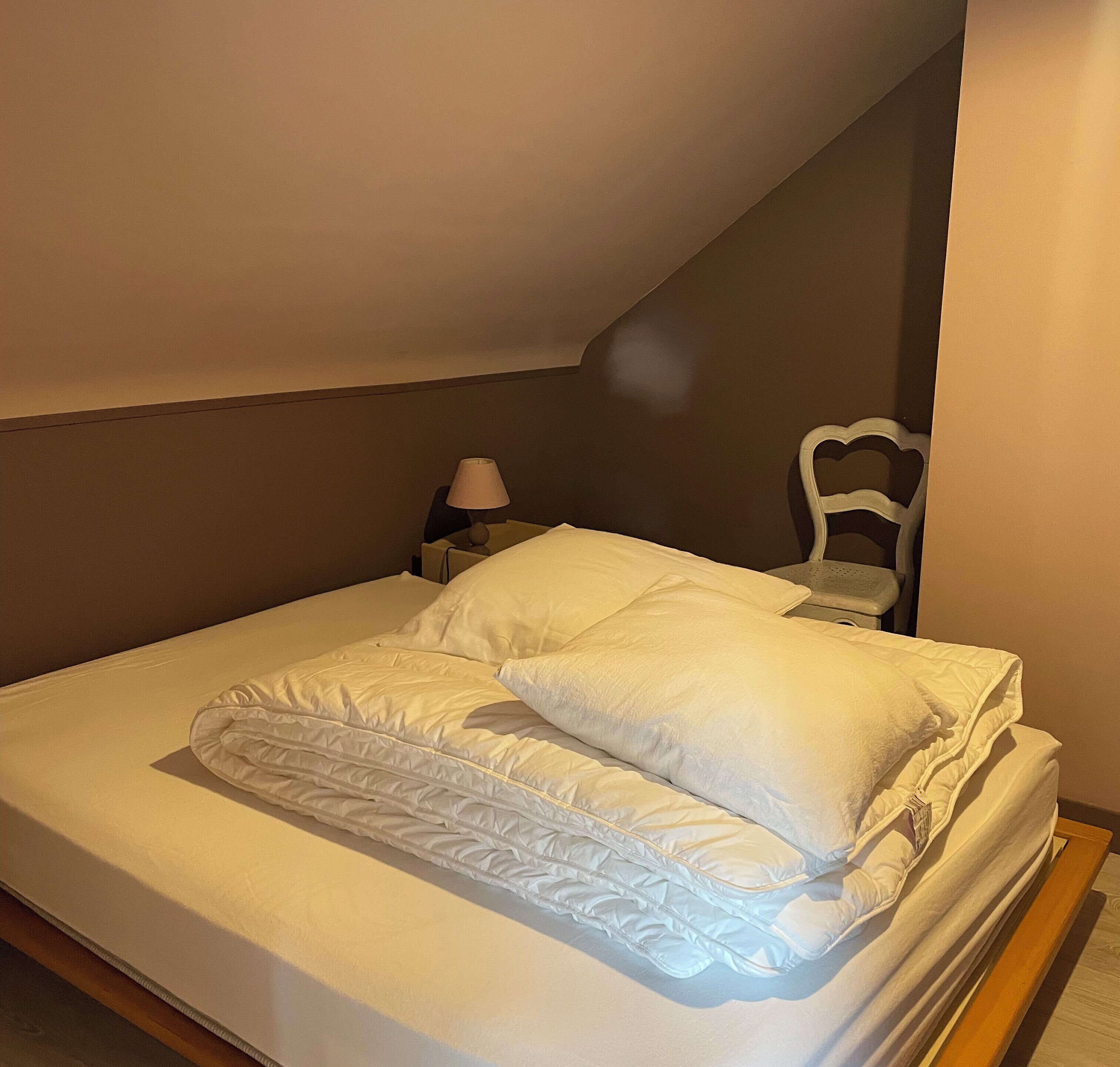 Una cama bien hecha con ropa blanca en una habitación pequeña y poco iluminada con techo inclinado.