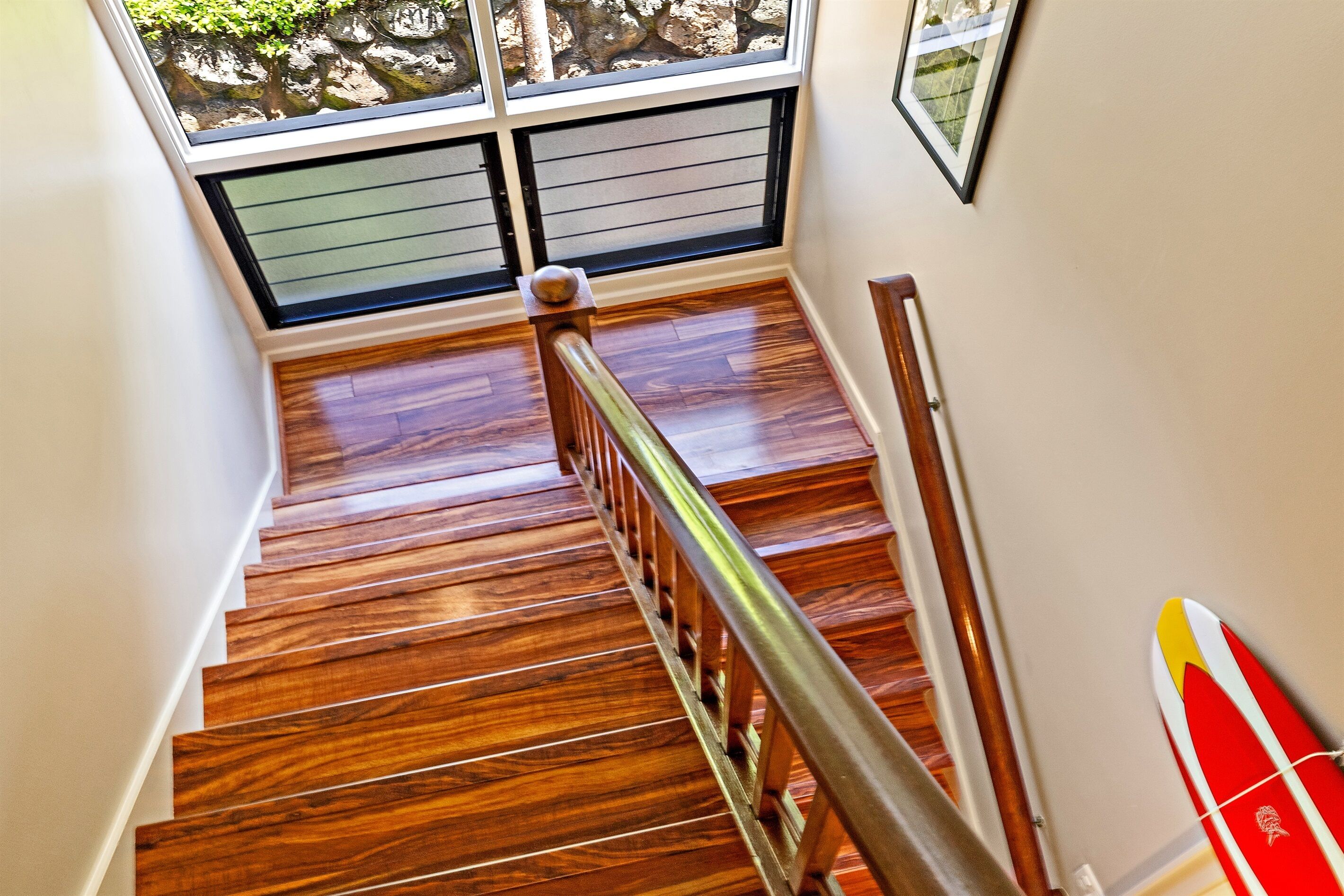 A wooden staircase with a metal handrail, leading to a room with a large window and a surfboard leaning against the wall.