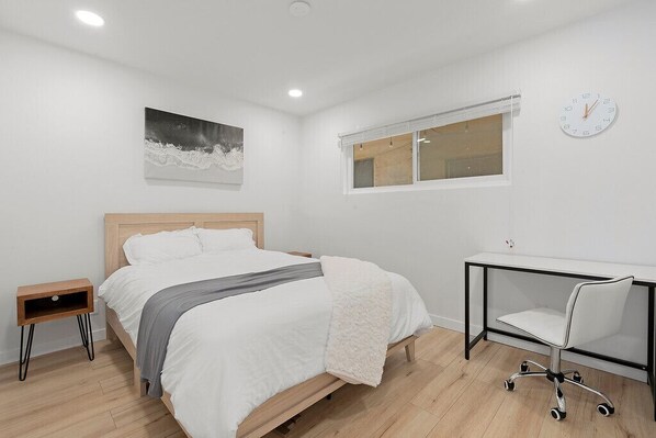 A bedroom with a bed, bedside table, desk, chair, and a clock on the wall.