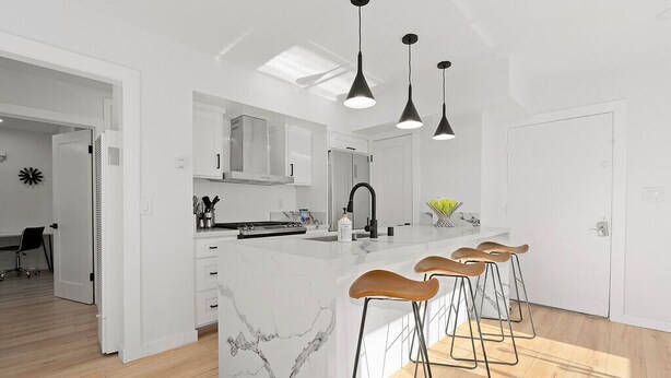 Una cocina moderna con una isla de mármol, taburetes de bar, iluminación colgante y vistas al área de comedor.