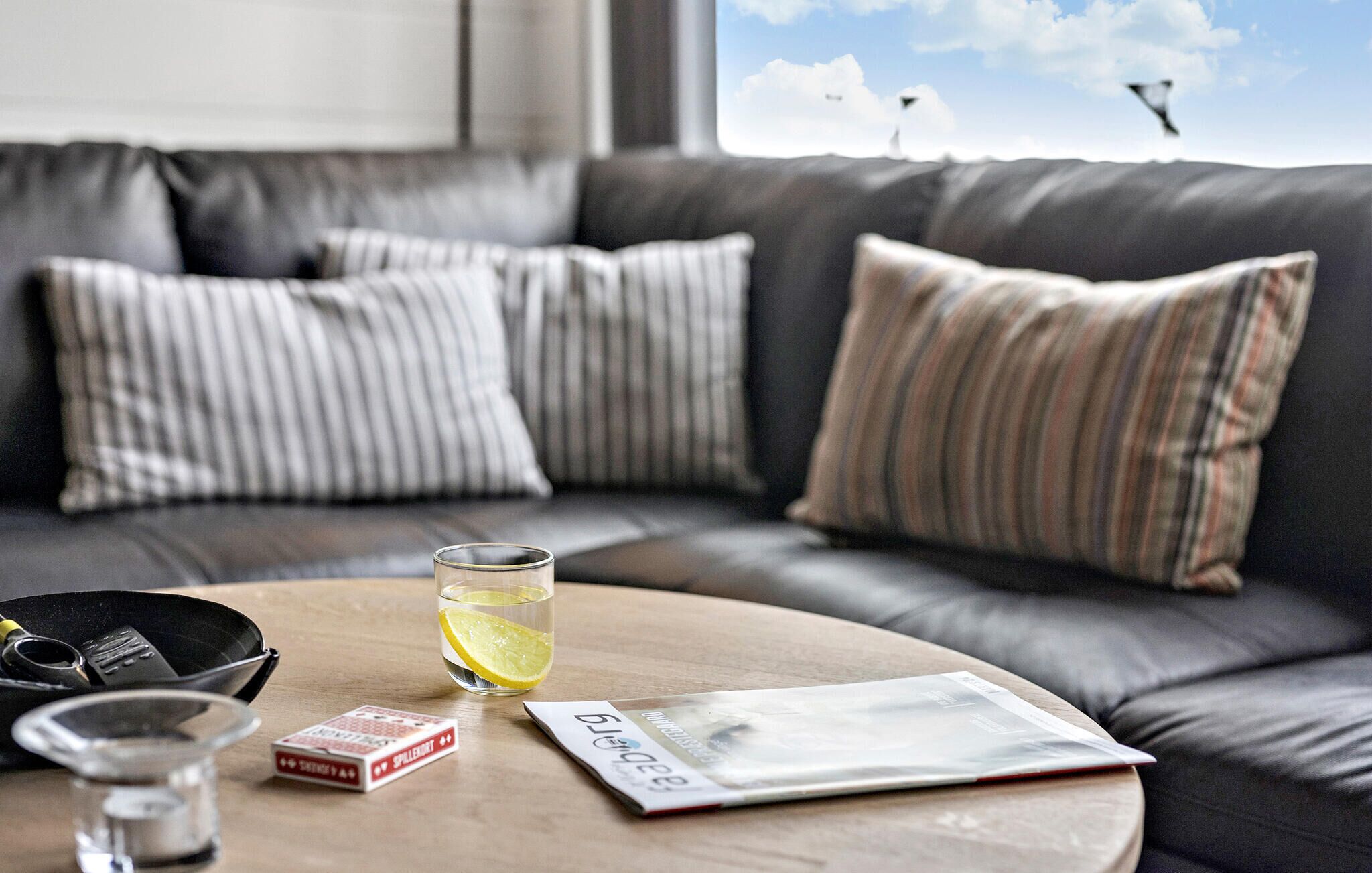 A modern living room with a leather sofa, striped pillows, a round coffee table, a glass of water with a lemon slice, a magazine, and a deck of playing cards.
