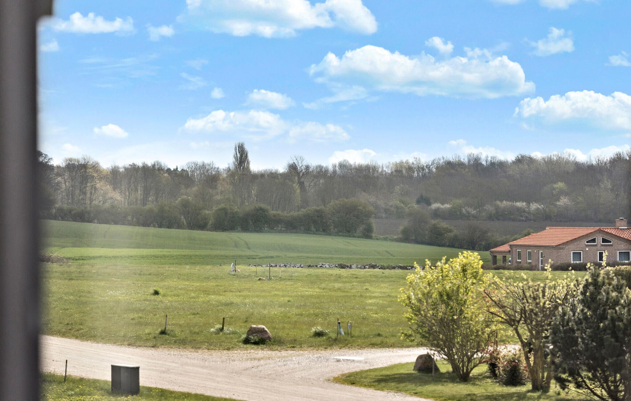 A rural landscape with a dirt road, green fields, a stone wall, and a brick building with a red roof.