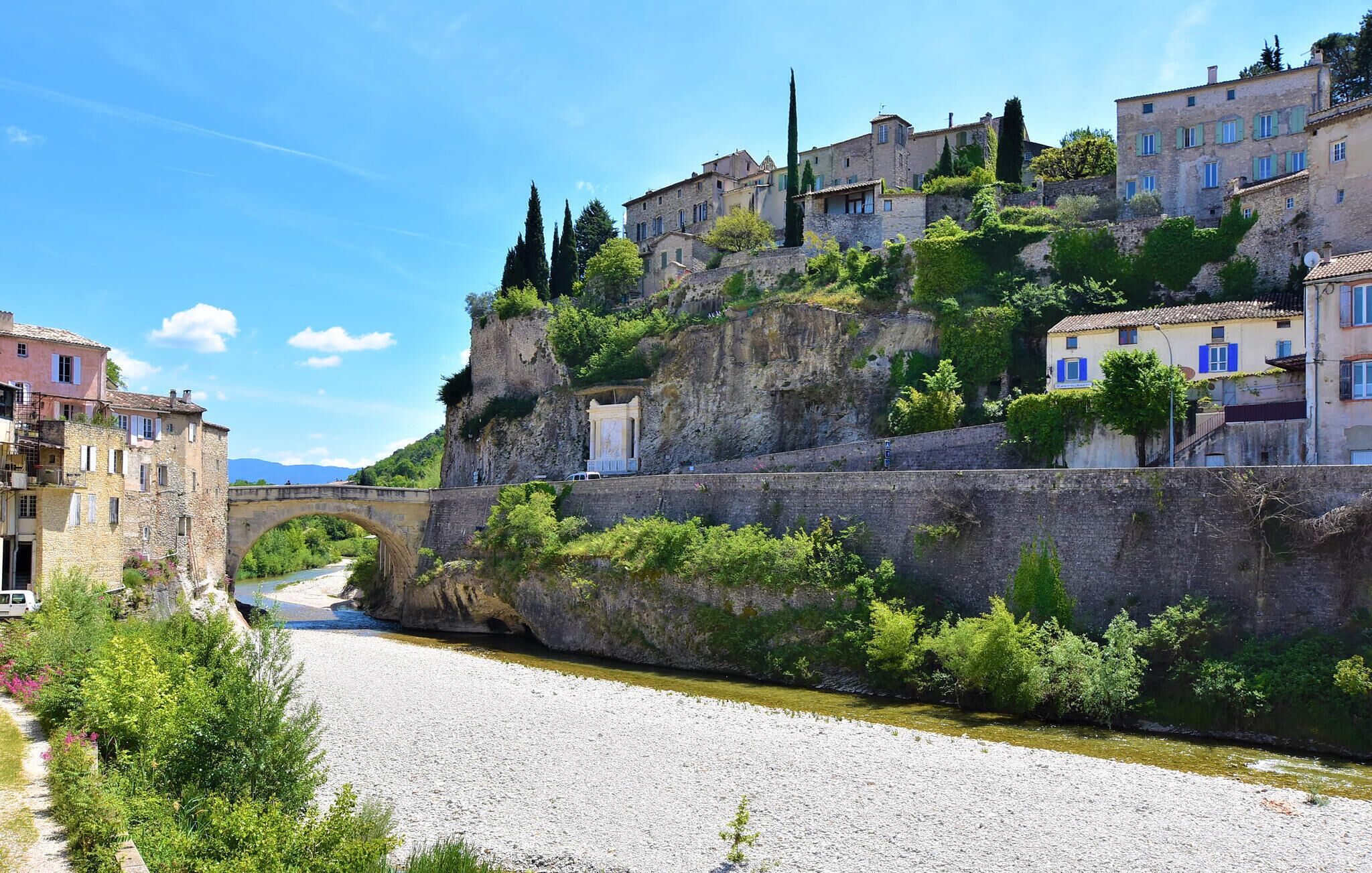 Un pont de pierre historique enjambant une rivière, avec des bâtiments sur une colline et un ciel bleu dégagé.