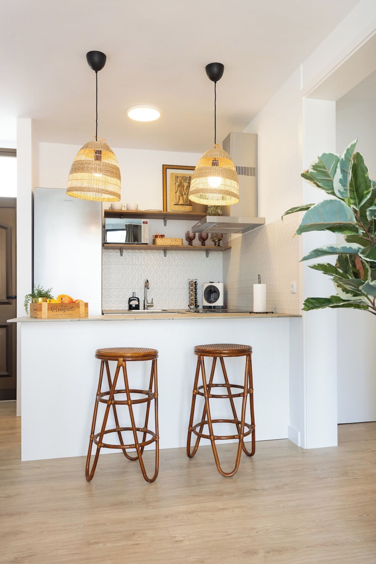 A modern kitchen with two hanging pendant lights, a built-in island with stools, and a large potted plant.