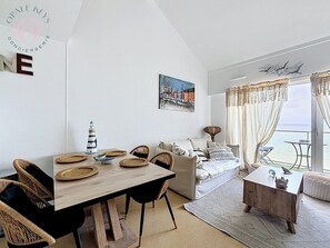 A modern living room with a wooden dining table, wicker chairs, a sofa, and a view of the sea through large windows. - Entre Ciel et Mer - Vue Mer - 3 CH & Parking (Bray-Dunes)