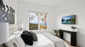 A bedroom with a bed, a window with a view of trees and mountains, a TV on a stand, and a bedside table with a plant.