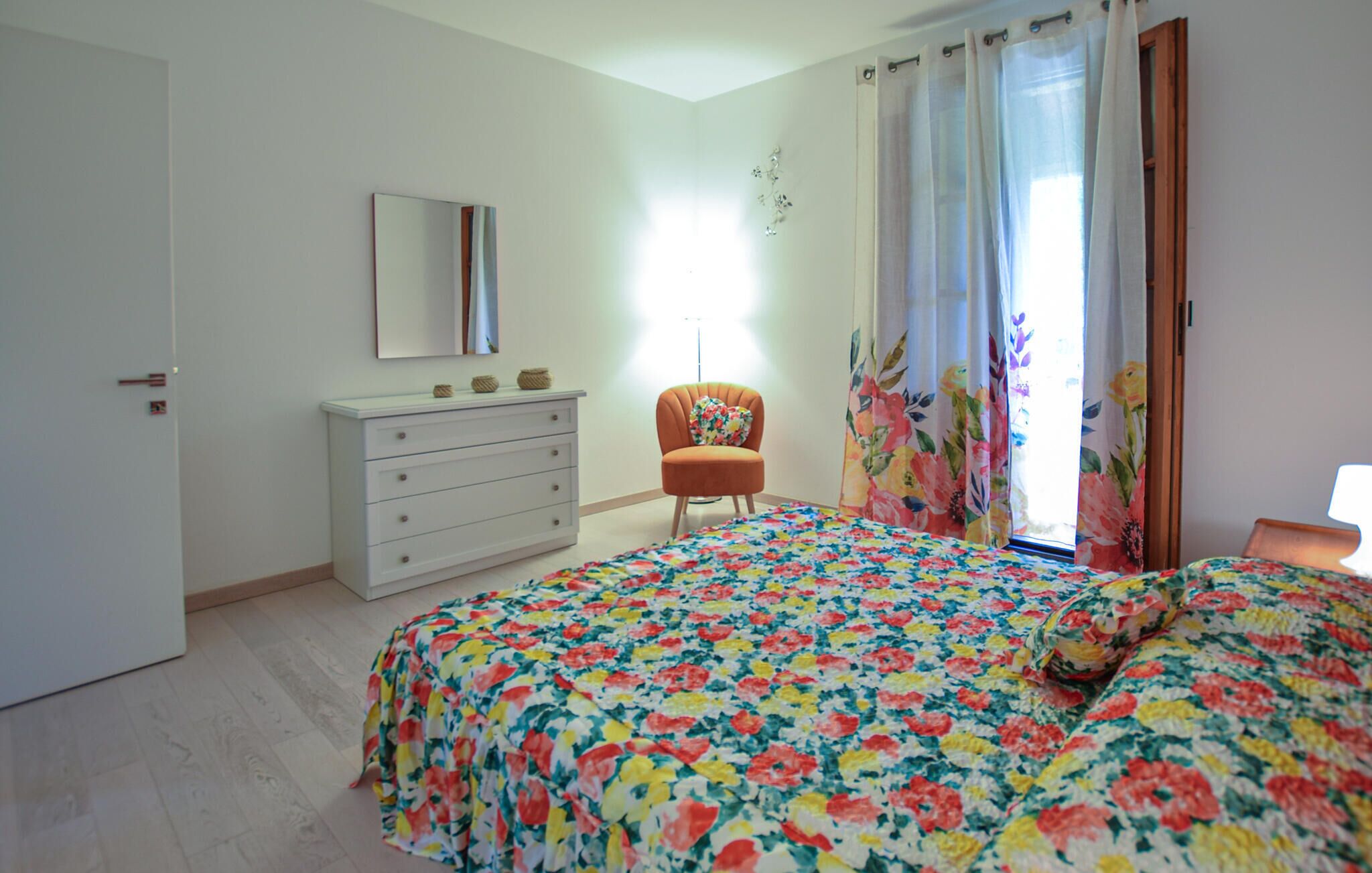 A bedroom with a floral bedspread, a white dresser, an orange armchair, and a window with floral curtains.