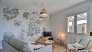 A modern living room with a large floral wall mural, a wooden coffee table, a grey sofa, a chair, a TV, and a window with a view of the outside. - Le Rocher - Trelivan (TRELIVAN)