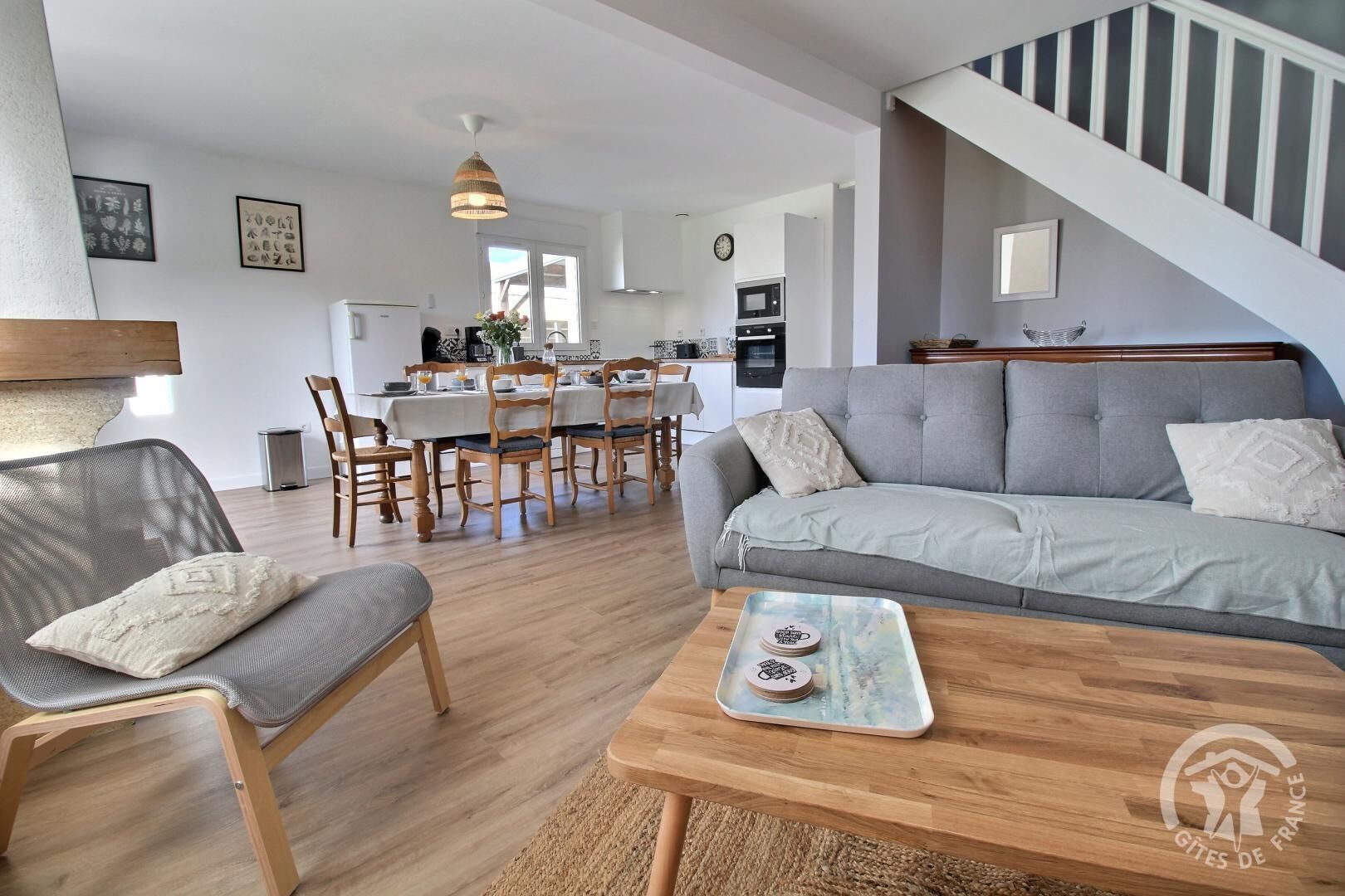 A modern living room with a grey sofa, wooden dining table, and chairs. A kitchen is visible in the background.