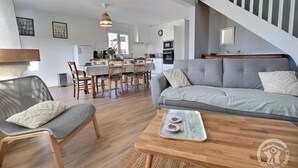 A modern living room with a grey sofa, wooden dining table, and chairs. A kitchen is visible in the background. - Le Rocher - Trelivan (TRELIVAN)