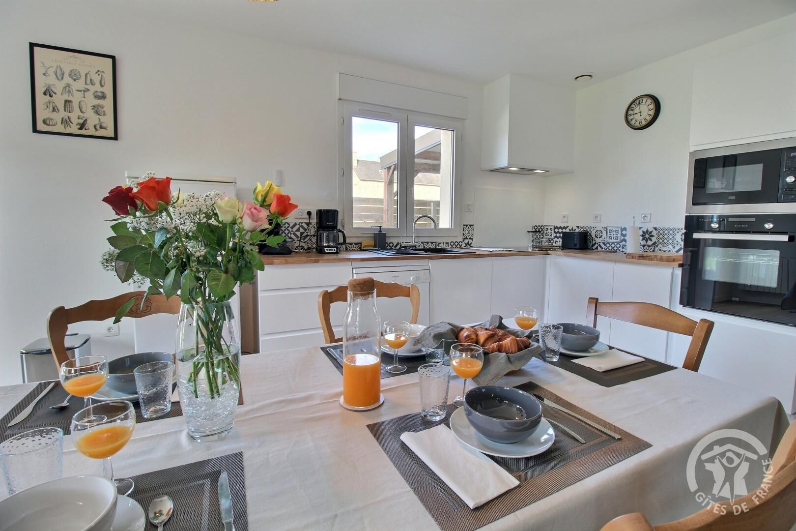 A modern kitchen with white cabinets, a coffee machine, and a dining area set for breakfast with orange juice and croissants.