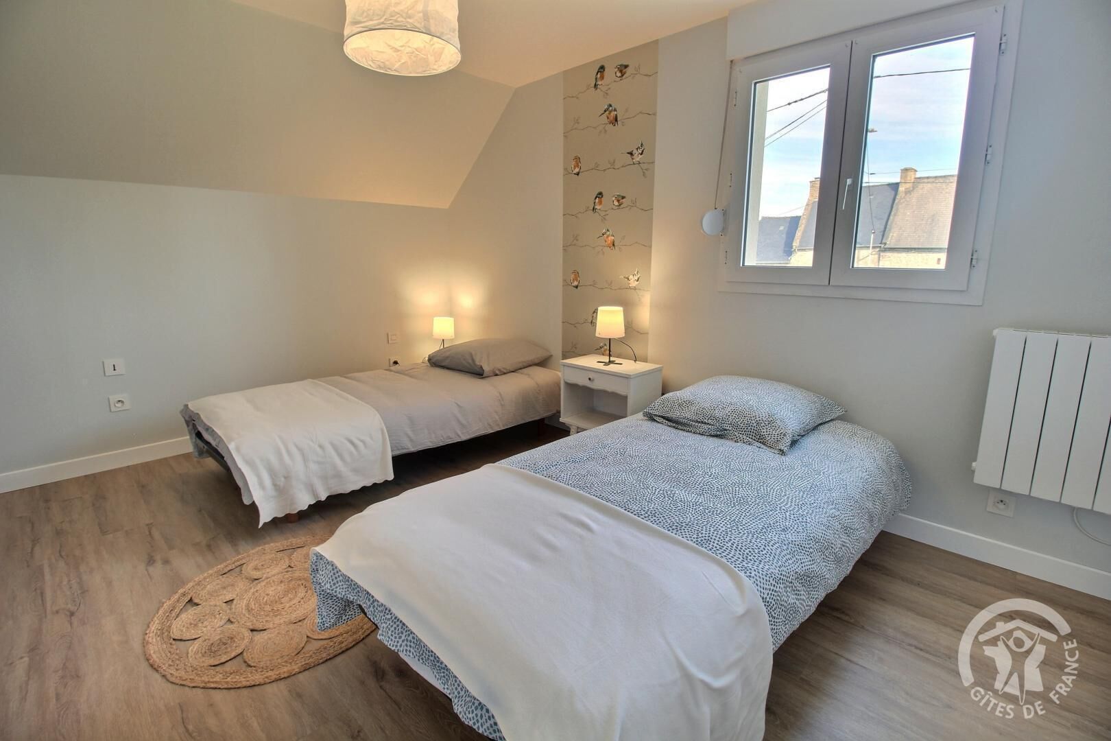 A bedroom with two beds, a wooden floor, a wall with a bird pattern, a window, and a ceiling light.