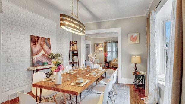 Un coin repas avec une longue table en bois dressée pour six personnes, des chaises blanches, un luminaire suspendu et un mur en briques.