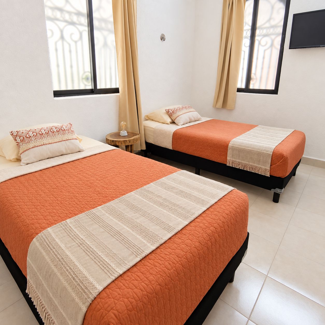 A hotel room with two beds, each with an orange quilt and a white throw. There are curtains on the windows, and a flat-screen TV mounted on the wall.