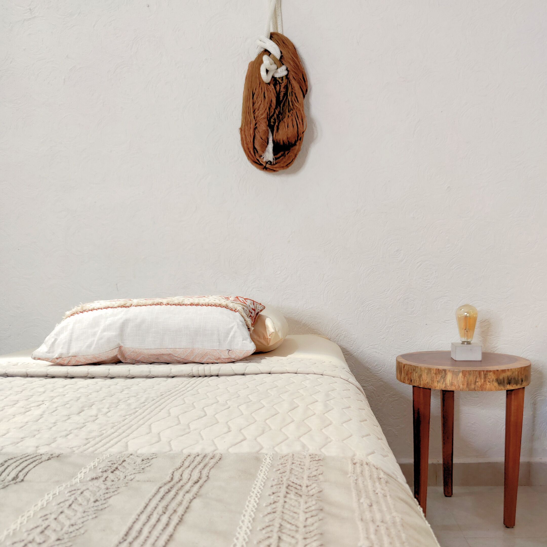 A bedroom with a bed, pillows, a hanging decoration, and a small wooden table with a lamp.