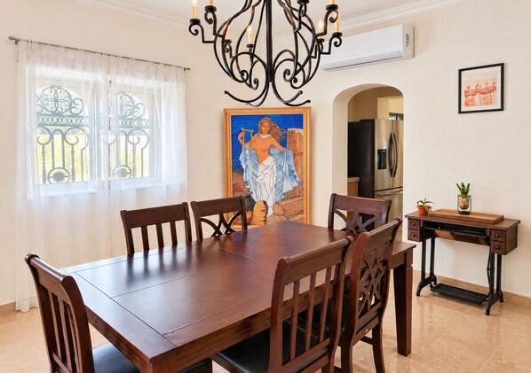 A dining area with a wooden table and chairs, a chandelier, a painting, and a kitchen in the background.