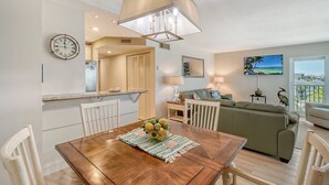 A dining area with a wooden table, chairs, and a bowl of fruit. There is a kitchen area with a refrigerator and a clock on the wall. A living room with a sofa and a painting is visible in the background. - Beachfront Condo - 2-Bedroom - Luxury - Directly on Crescent Beach (Siesta Key)