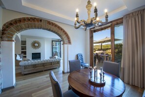 A dining area with a round wooden table, a chandelier, and a brick archway leading to a living room with a sofa and television.