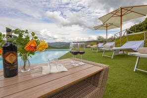 A wooden table with a bottle of Comignol, two glasses of red wine, and a vase with roses by a poolside lounge area with white chairs and umbrellas. - MyExclusiveItaly - Villa Chianti with pool near the vineyard (Greve in Chianti)