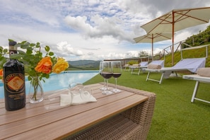 Une table en bois, une bouteille de Comignol, deux verres de vin rouge et un vase de roses, le tout placé près d’un coin salon au bord de la piscine, avec des chaises et des parasols blancs.