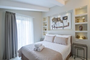 Une chambre d’hôtel avec un grand lit, une fenêtre avec des rideaux, une étagère fixée au mur ornée d’objets décoratifs et une petite table.