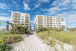 Un hôtel situé en bord de mer, doté de nombreux balcons, d’un chemin de sable menant à l’entrée et surplombant un ciel d’un bleu éclatant.