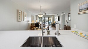 A modern kitchen with a central island, stainless steel sinks, and a dining area with wicker chairs and a chandelier. - Seashore Splendor with Ocean views, Heated Pool & Tennis Court (Cocoa Beach)