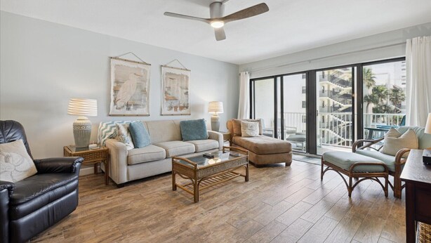 A spacious living room with a sofa, armchairs, and a coffee table. There are two wall-mounted lamps, a ceiling fan, and large windows offering a view of the outside. - Seashore Splendor with Ocean views, Heated Pool & Tennis Court (Cocoa Beach)