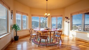 A dining area with a round table, chairs, a chandelier, and large windows with a view of a forested landscape.