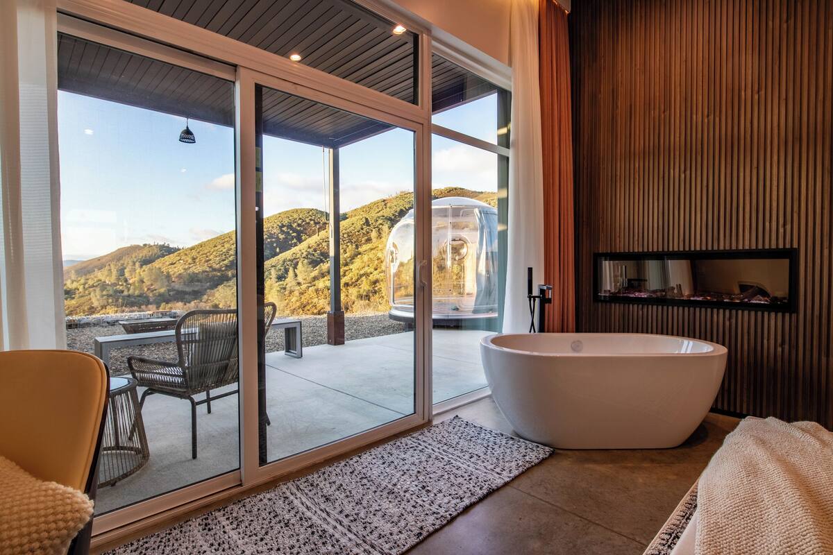 A modern bathroom with a large bathtub, a glass-enclosed shower, and a view of a hillside.