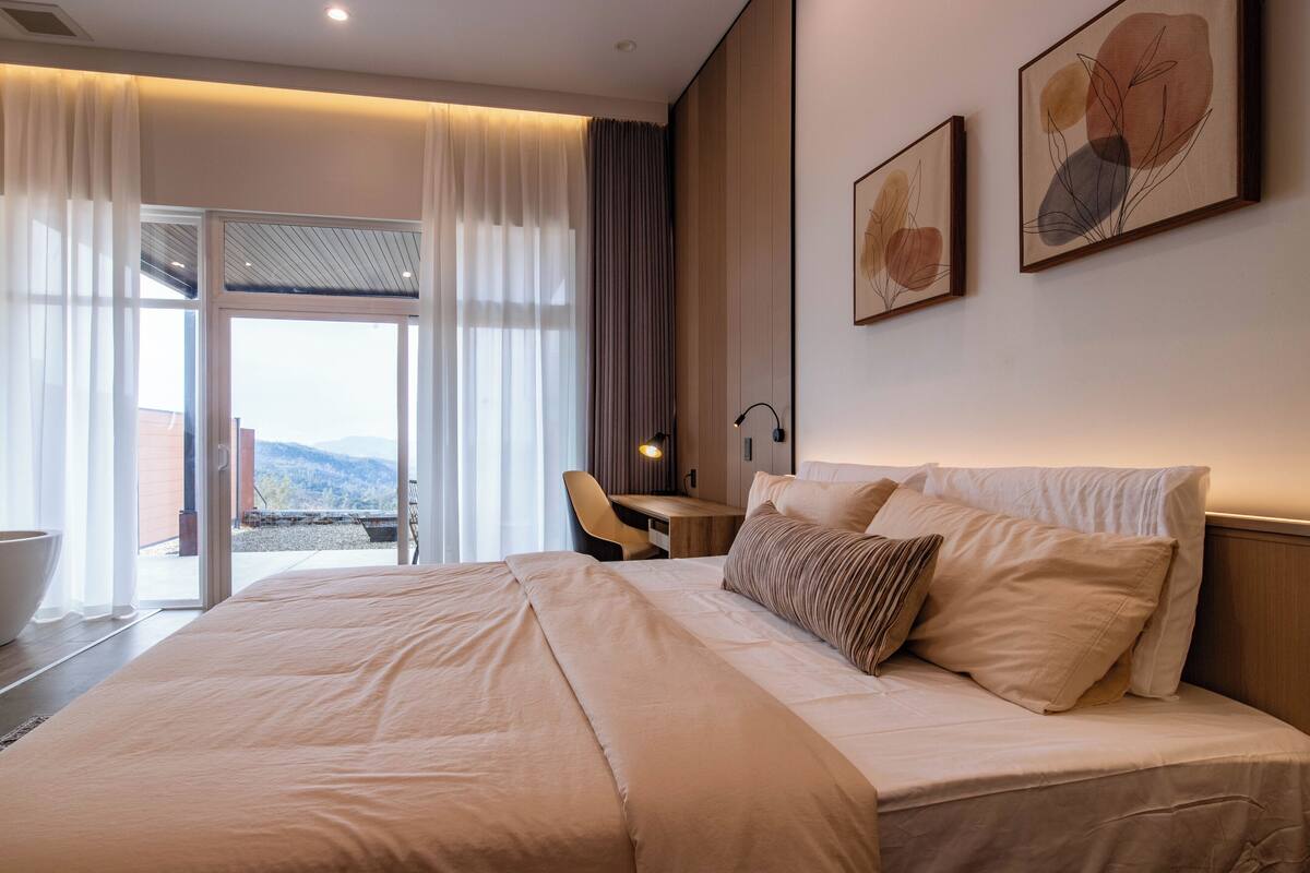 A hotel room with a large bed, a desk, and a chair. There is a window with curtains and a view of the outside.