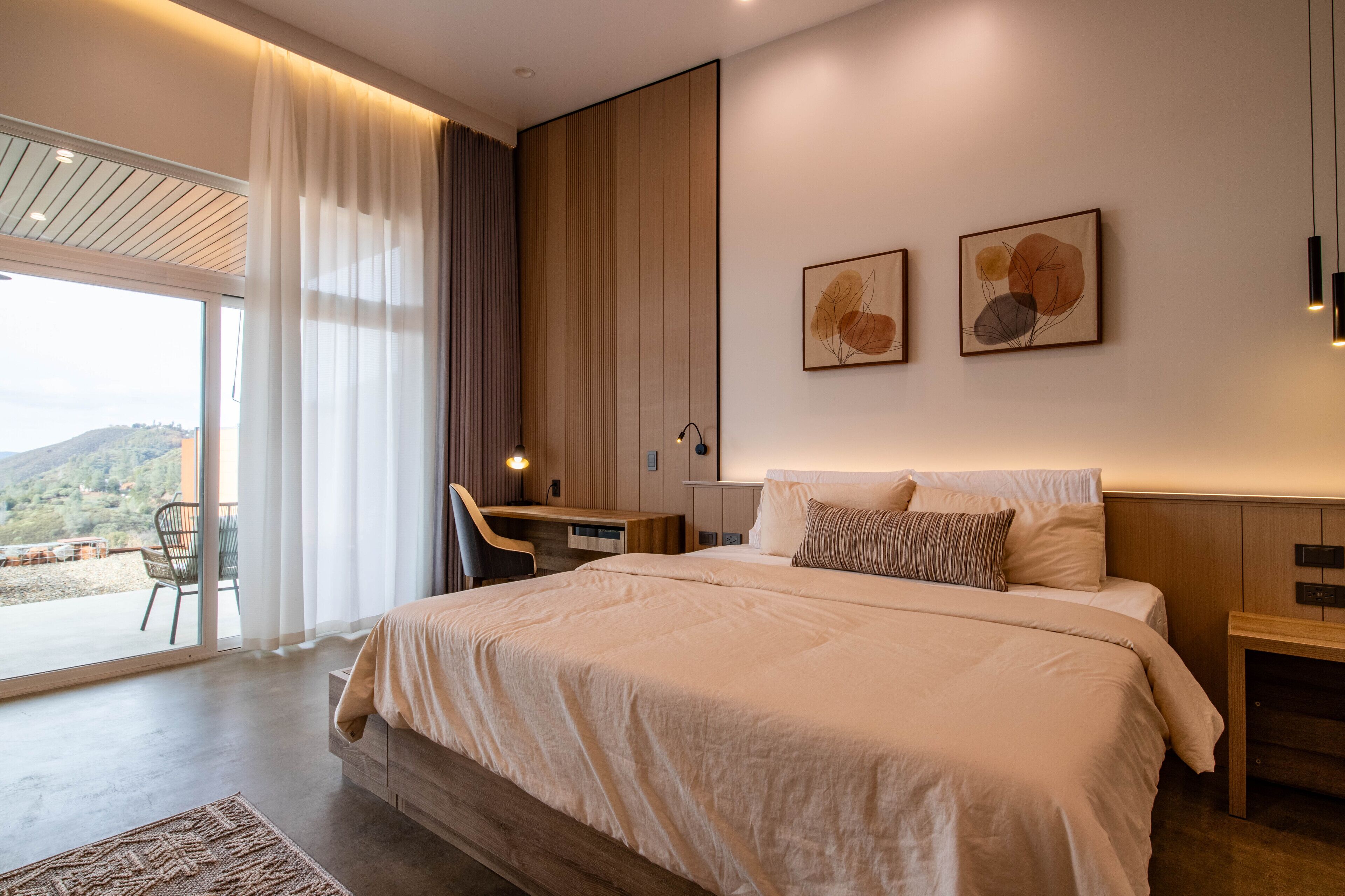 Une chambre moderne avec un grand lit, un bureau et une chaise, une grande fenêtre avec des rideaux et deux œuvres d’art accrochées au mur.