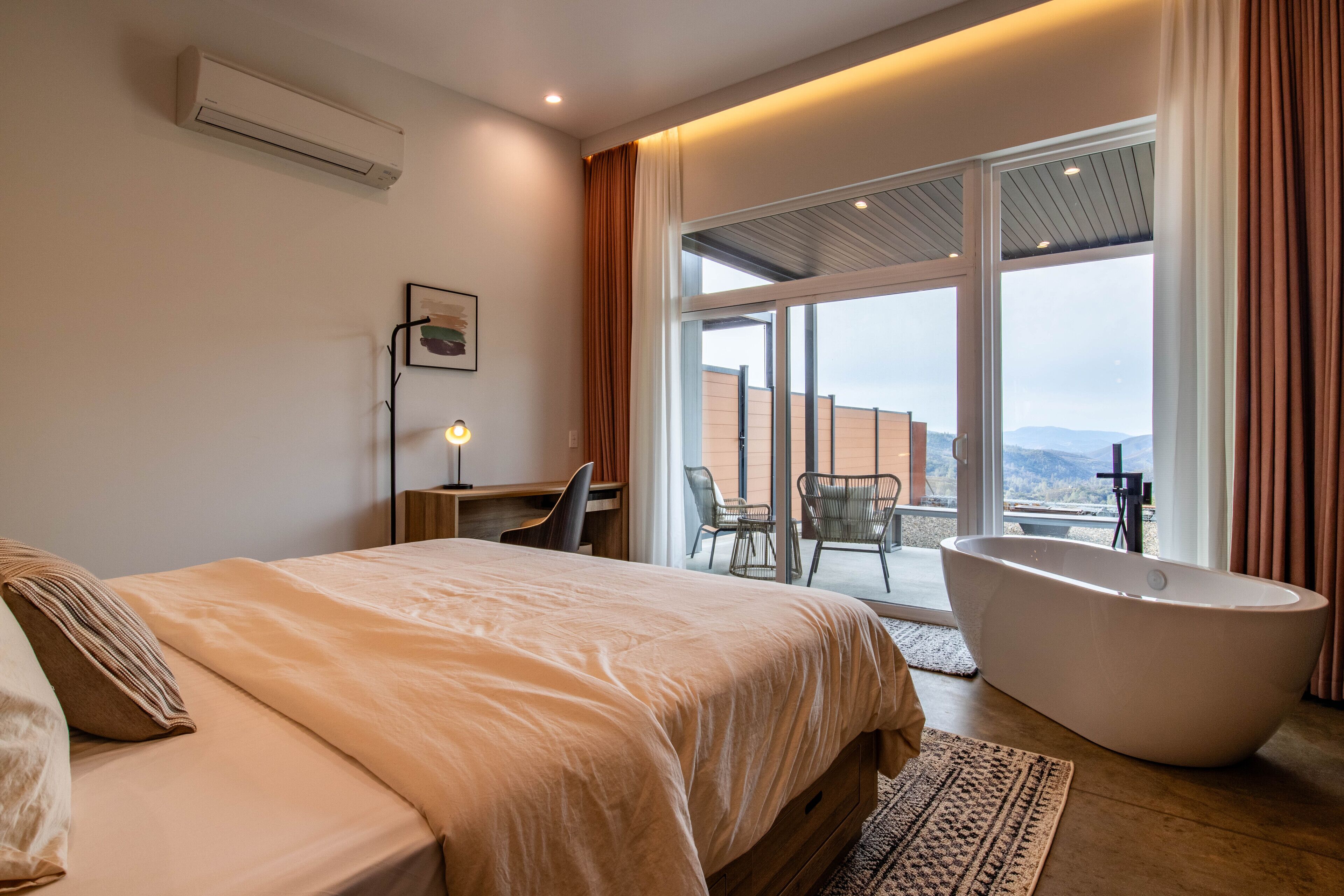 A modern hotel room with a large bed, a sitting area with a desk and chair, a bathtub, and a balcony with a view of the mountains.