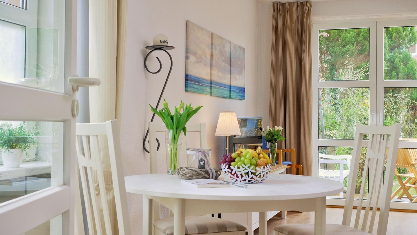 A dining area with a white table, chairs, a bowl of fruit, a lamp, a painting, and a view of greenery outside.