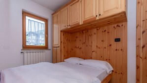 A bedroom with a wooden headboard, a window with a lace curtain, and a bed with white bedding. - Casa Stella 4 (Falcade)