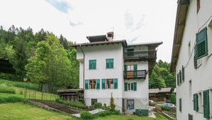 A two-story building with green shutters and a balcony, surrounded by a grassy area and trees. - Casa Silva (Falcade)