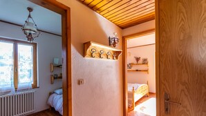 A bedroom with a wooden ceiling, a bed with a wooden headboard, a radiator, a window with lace curtains, and a wall-mounted shelf with hooks.