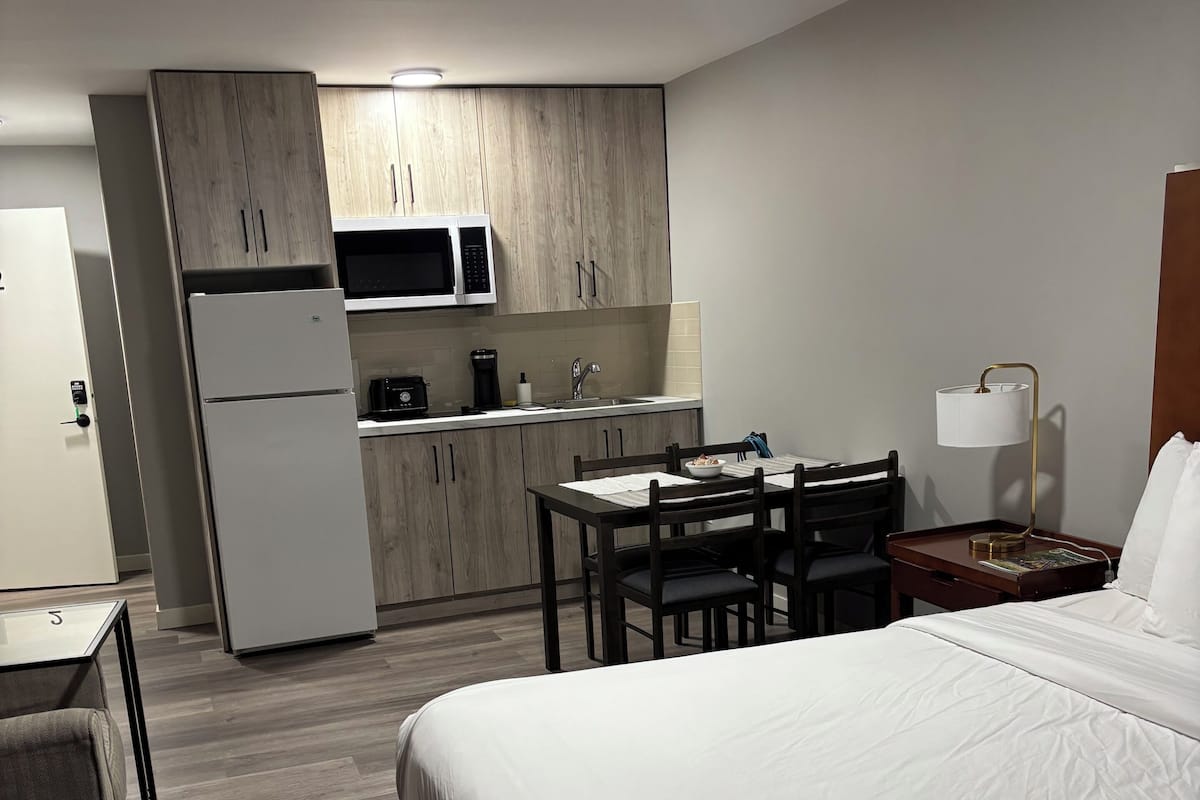 Et hotelværelse med seng, skrivebord med stol, lille bord med stol samt et tekøkken med køleskab, mikroovn og vask.