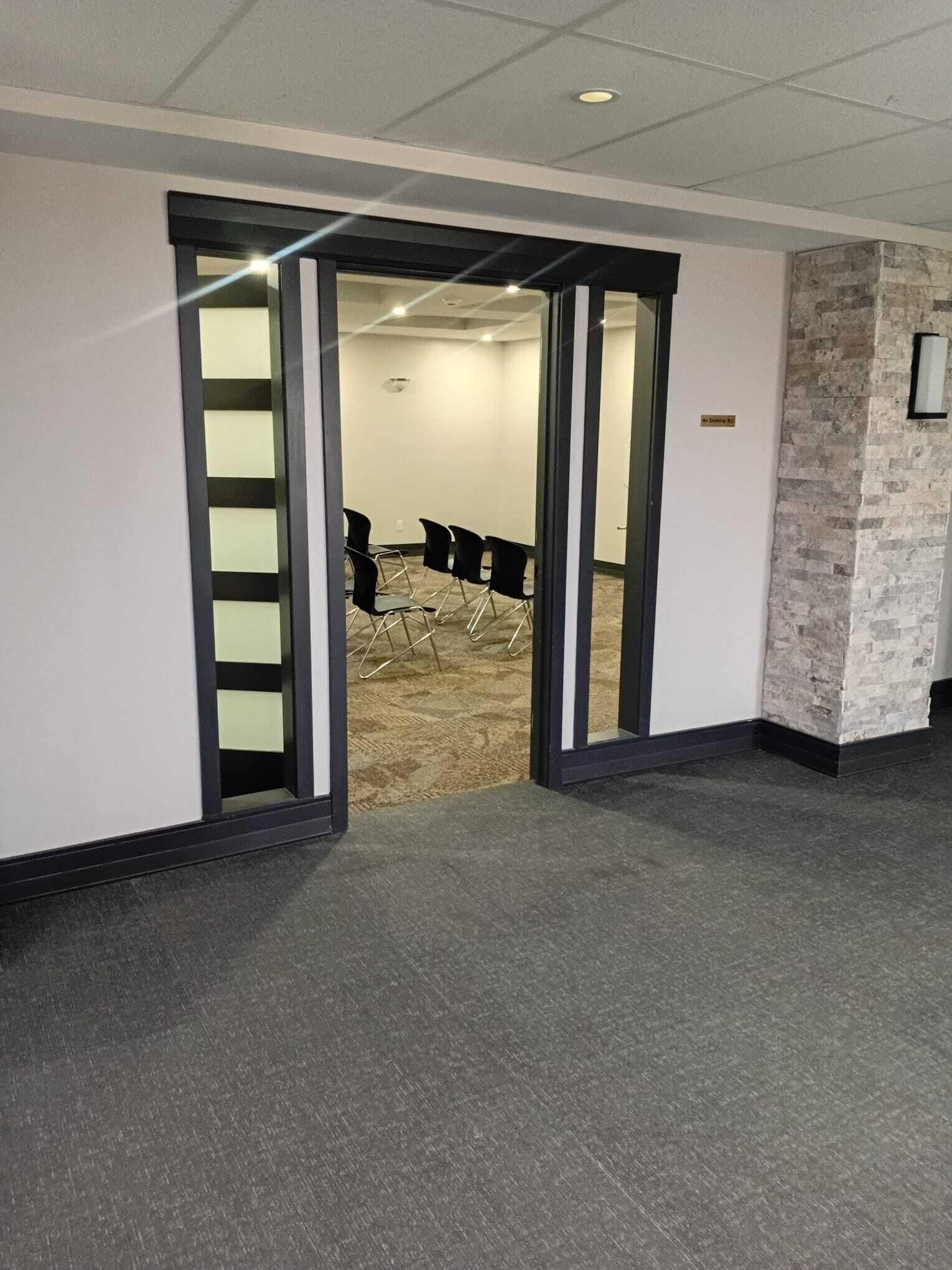 An interior corridor with a carpeted floor, a brick accent wall, and a door leading to a room with chairs.