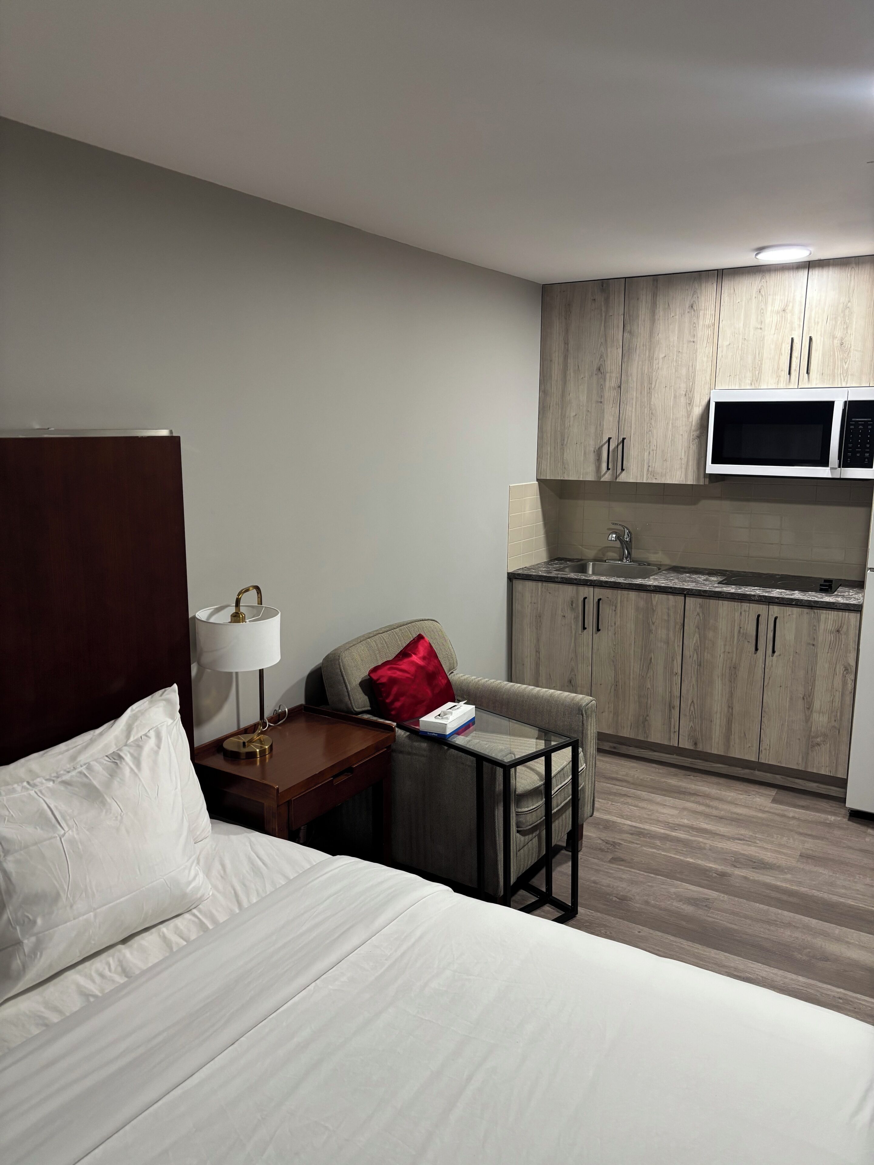 A hotel room with a bed, a bedside table with a lamp, a chair, a small table, a microwave, and a kitchenette.