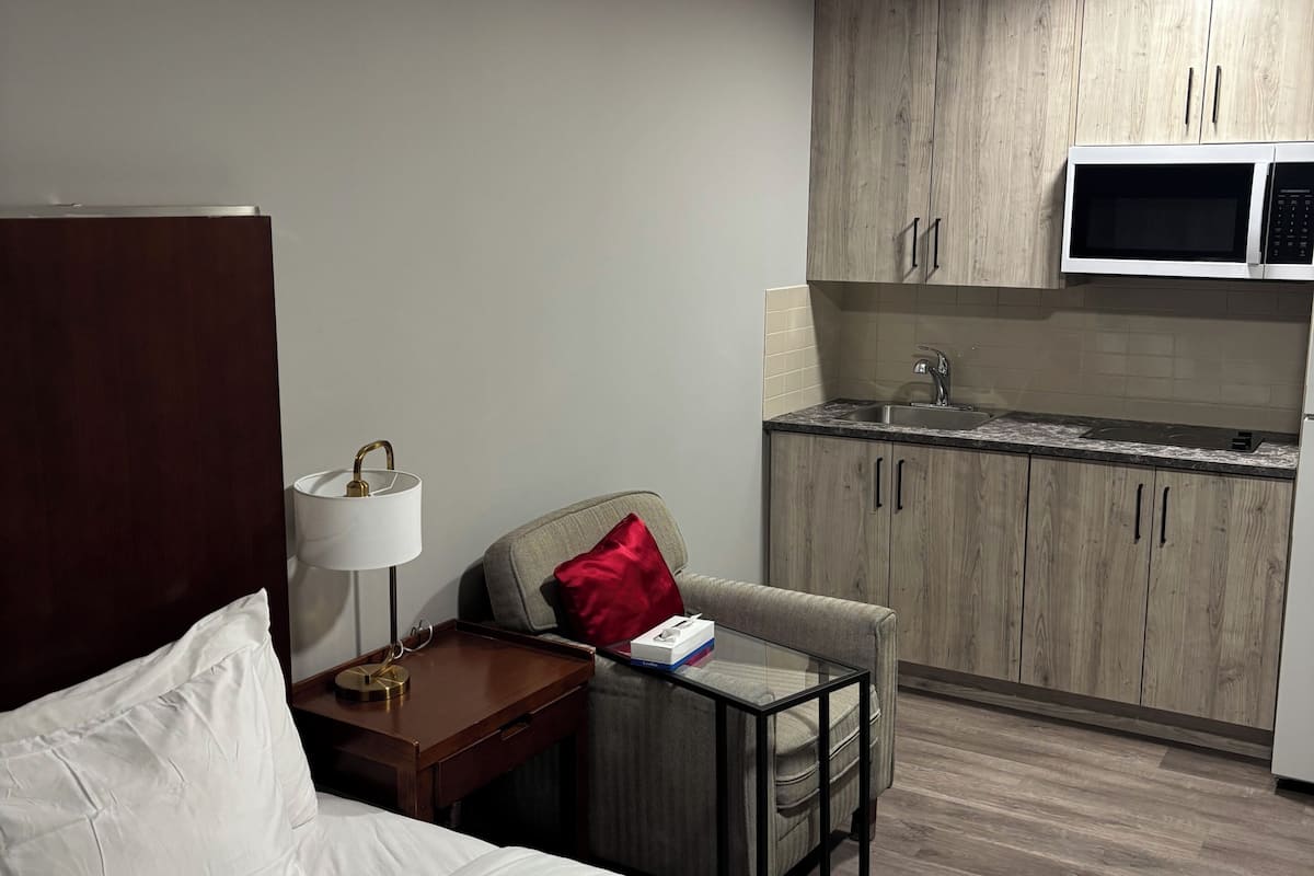 A hotel room with a bed, a bedside table with a lamp, a chair, a small table, a microwave, and a kitchenette.