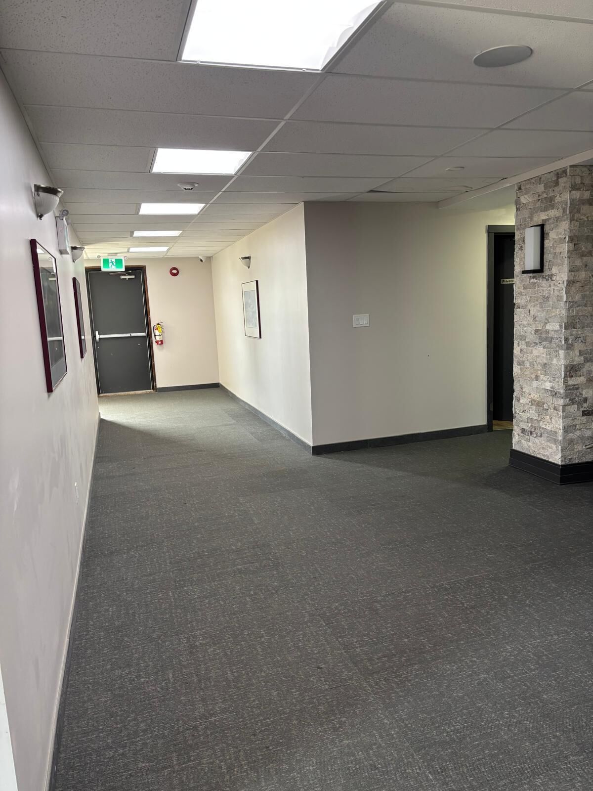 Un pasillo con alfombra gris, paredes blancas y una pared de acento de piedra. Hay cuadros enmarcados en la pared, un extintor y una puerta con un cartel.