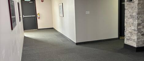 Un pasillo con alfombra gris, paredes blancas y una pared de acento de piedra. Hay cuadros enmarcados en la pared, un extintor y una puerta con un cartel.