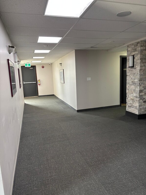 Un couloir avec un tapis gris, des murs blancs et un mur d’accent en pierre. On y trouve des tableaux encadrés, un extincteur et une porte avec une pancarte.
