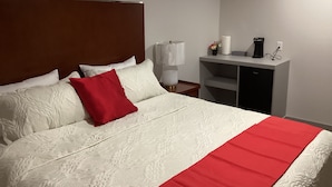 A neatly made bed with a red accent pillow, a bedside lamp, and a small table with a coffee maker and a vase of flowers. - Bethel Hotel Elk Point (Elk Point)