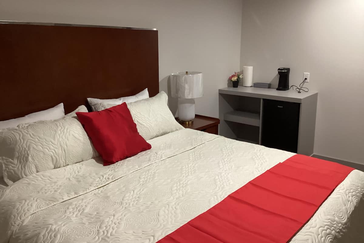 A neatly made bed with a red accent pillow, a bedside lamp, and a small table with a coffee maker and a vase of flowers.